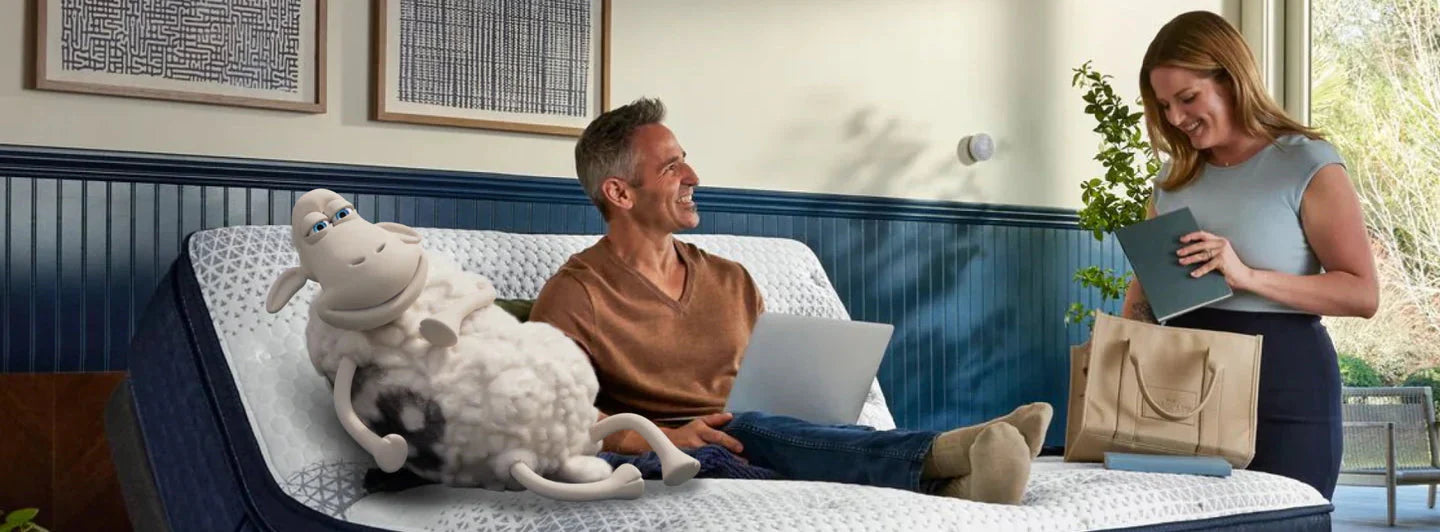 Man relaxing on a Serta adjustable bed frame with a laptop, while a Serta sheep lounges beside him, showcasing customizable comfort and modern sleep technology.