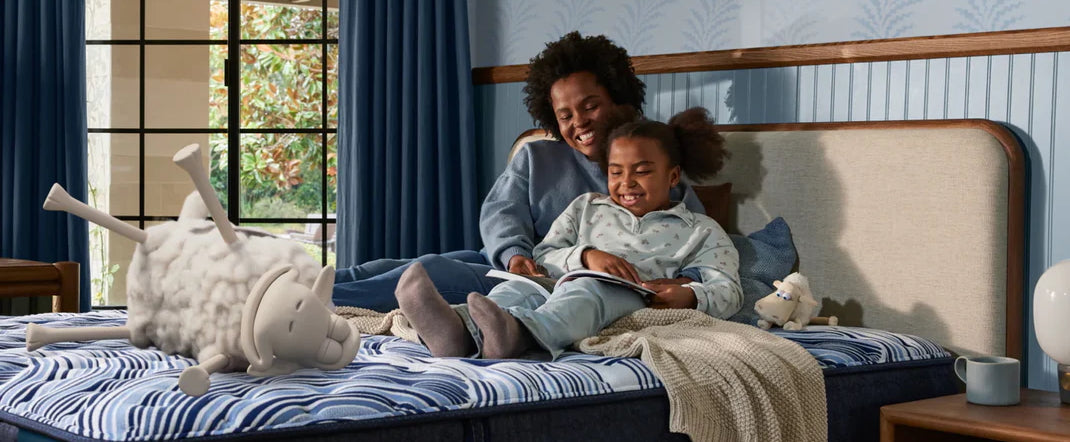 Woman and daughter laying on a twin xl mattress while reading a book and cuddling. A plush stuffie and serta sheep also lay on the bed.