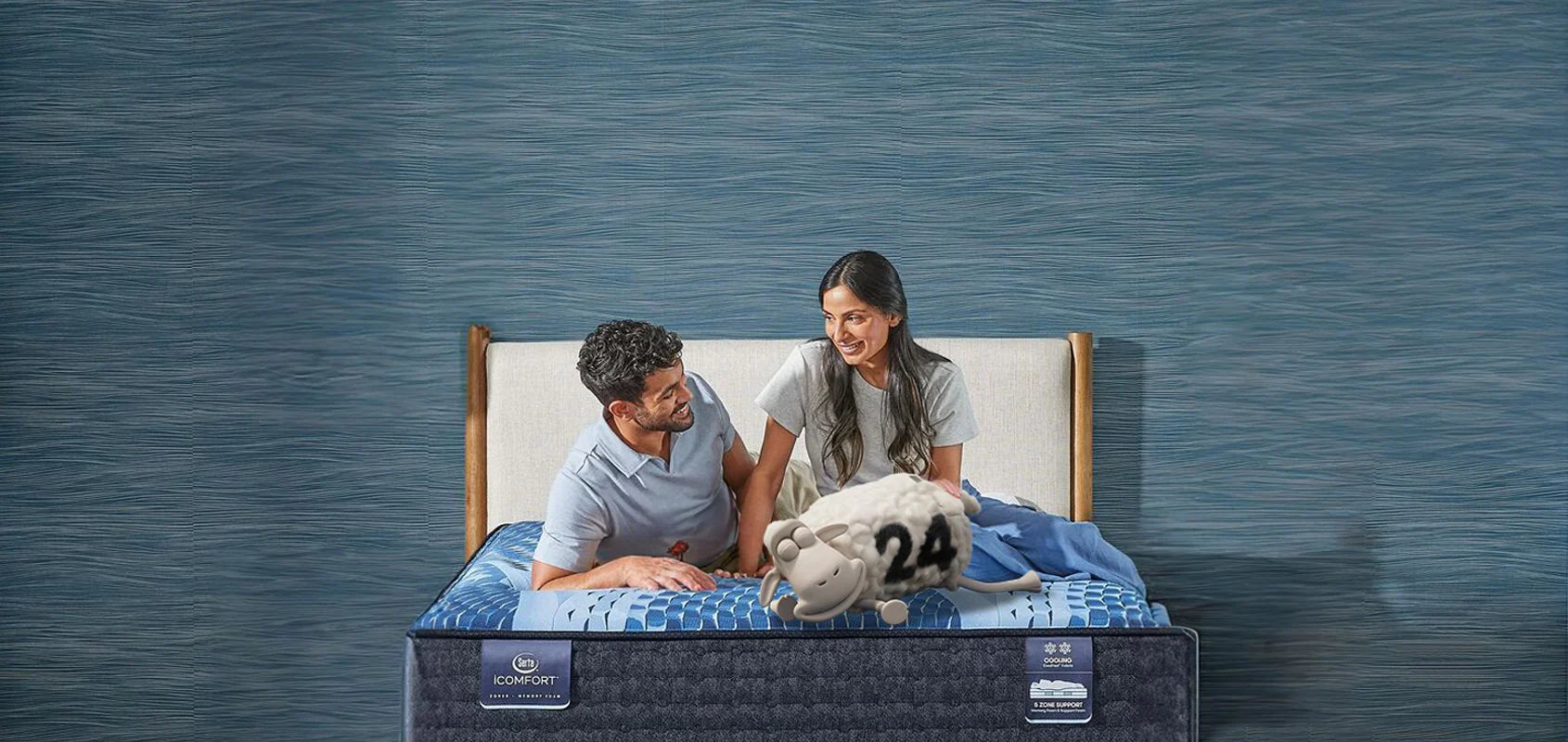 Smiling couple sits on a Serta iComfort mattress, featuring cooling technology and zoned support, with a relaxed Serta sheep lying on the bed.