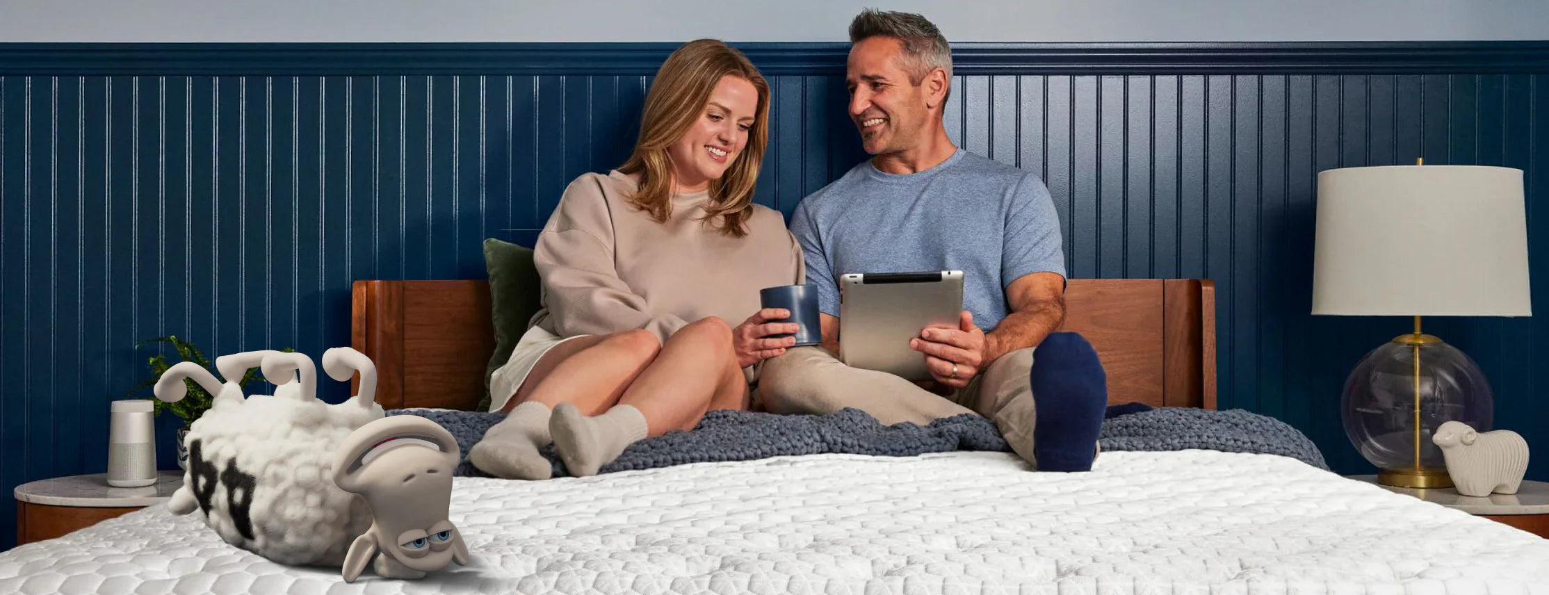 Smiling couple sits on a Serta Queen mattress in a cozy bedroom, with a Serta sheep playfully lounging nearby, emphasizing spacious comfort for couples and solo sleepers.