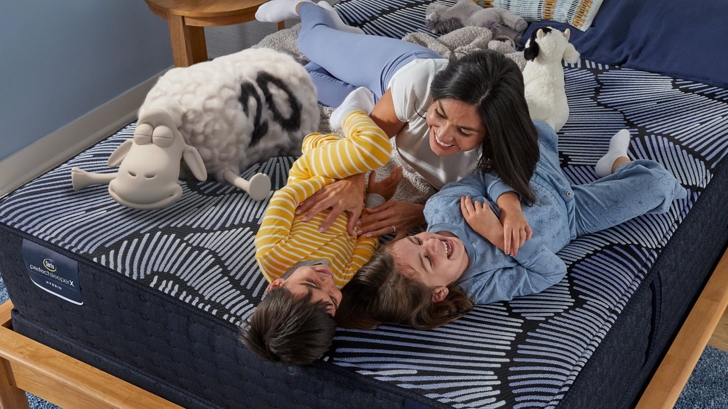 Mom tickling two laughing kids on a Serta mattress while the Serta sheep smiles next to them